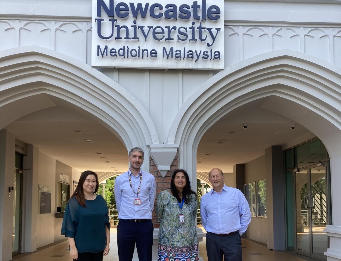 Four people in front of a grand entrance with a sign saying Newcastle University Medicine Malaysia