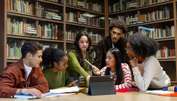 A group of diverse students in a library collaborating around a laptop