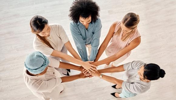 A group of five diverse women stand in a semi circle with their arms outstretched and hands in the middle