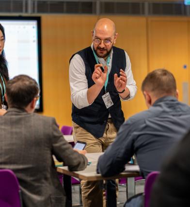 A group of participants seated around tables in a conference room while two facilitators stand and speak to them. A large presentation screen is visible in the background, setting appears to be an interactive workshop or breakout session.